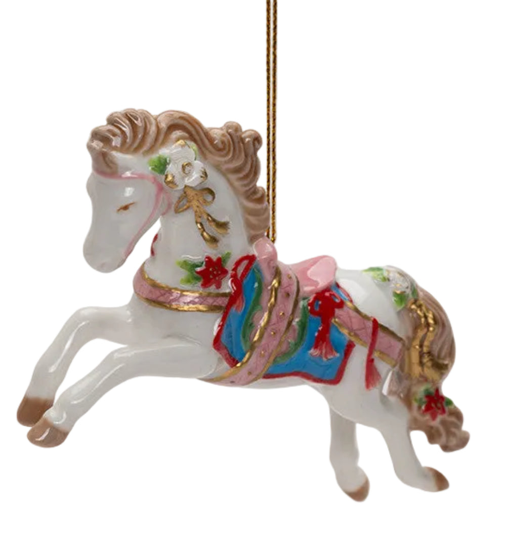 A white porcelain Christmas ornament shaped like a rearing horse, decorated with a pink saddle, blue and red patterned blanket, green leaves, and gold accents, hanging from a gold string against a light gray background.
